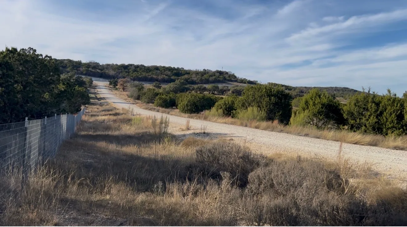 A gravel ranch road running along the fence line through the Texas Hill Country